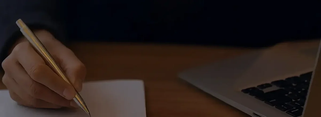 A hand holding a pen above a notebook, with a laptop partially visible.