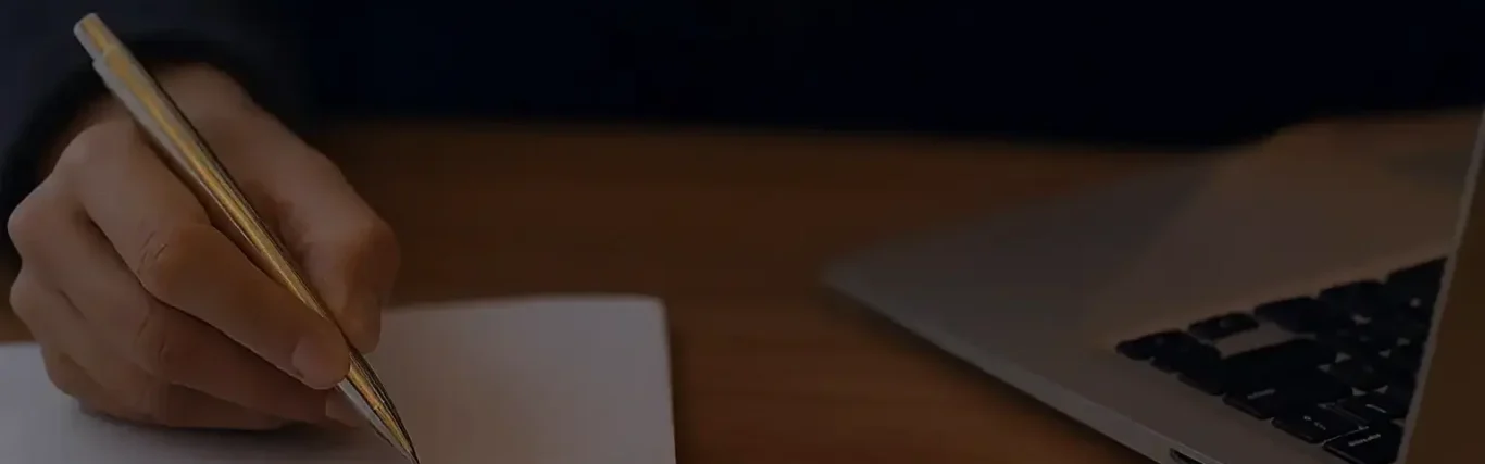 A hand holding a pencil writes on paper next to a laptop.